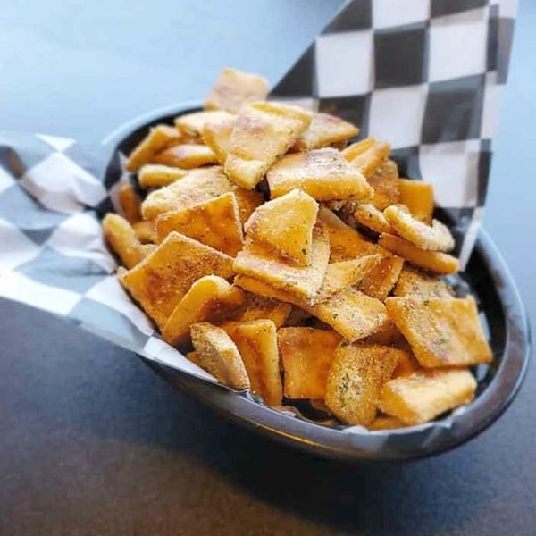 A black bowl filled with golden-brown pita chips, seasoned and lined with checkered paper.