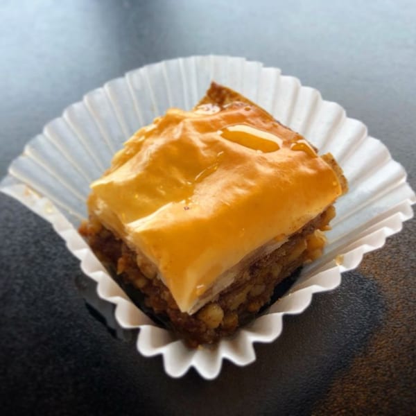 A golden-brown piece of baklava, drizzled with honey, sits in a white paper cup.