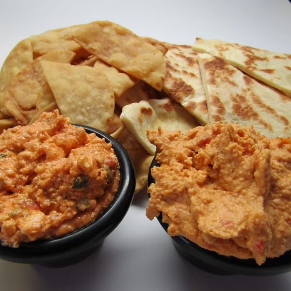Two bowls of orange Greek dip with pita bread and fried pita chips on a white surface.