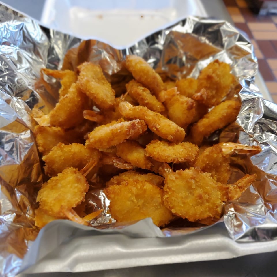 Golden-brown fried shrimp with tails on, served in a foil-lined takeout container.